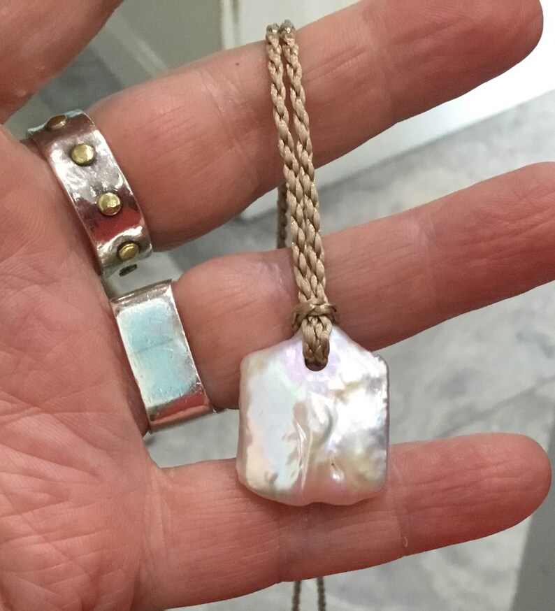 May include: A pendant featuring an irregular, iridescent white baroque pearl suspended from a braided tan cord. The pearl has a flat, rectangular shape with a soft lustre. Two silver rings are also visible, one with gold-toned studs.