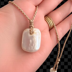 May include: A rectangular, iridescent white pearl pendant suspended from a thin, braided tan cord. The pendant features a smooth, polished surface with a soft lustre. The cord includes a small, round white bead at the end of the adjustable closure.