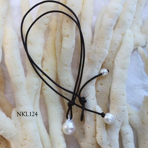 Puede incluir: Un collar de cord&oacute;n negro con tres perlas blancas. Una perla tiene forma de l&aacute;grima, las otras dos son redondas. El collar se muestra sobre un fondo texturizado blanquecino. El texto "NKL124" es visible.