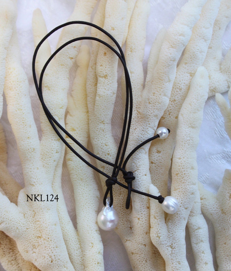 Puede incluir: Un collar de cord&oacute;n negro con tres perlas blancas. Una perla tiene forma de l&aacute;grima, las otras dos son redondas. El collar se muestra sobre un fondo texturizado blanquecino. El texto "NKL124" es visible.
