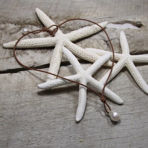 Puede incluir: Un collar de cuero marr&oacute;n con un solo colgante de perla blanca. El collar se exhibe sobre una superficie de madera con varias estrellas de mar blancas.