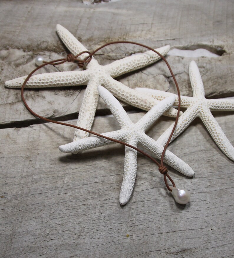 Puede incluir: Un collar de cuero marr&oacute;n con un solo colgante de perla blanca. El collar se exhibe sobre una superficie de madera con varias estrellas de mar blancas.