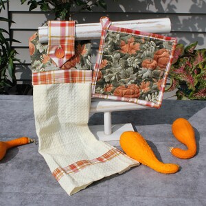 May include: Two fall-themed kitchen towels with a plaid pattern and a pumpkin and leaf design. The towels are hanging on a white stand with two orange gourds in front.