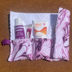 May include: A white and purple marbled fabric pouch with a zipper closure. The pouch contains a white bottle of body wash, a white tube of sunscreen lotion labeled "sport sunscreen lotion", and a pink razor.