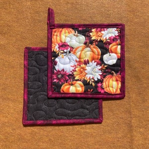 May include: Two black and burgundy quilted pot holders with a fall-themed print of pumpkins, flowers, and leaves.
