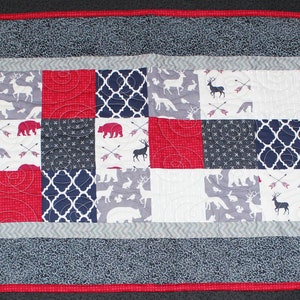 May include: A red, white, and blue patchwork table runner with a woodland theme. The runner features bears, deer, and arrows on a white background. The runner is bordered with a red and gray trim.