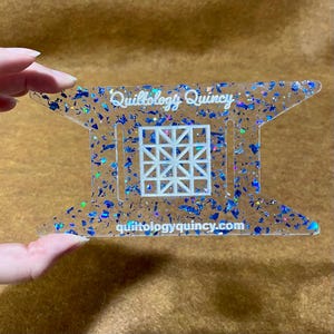 May include: A clear acrylic quilting template with blue and iridescent glitter. The template features a geometric design and the text "Quiltology Quincy" and "quiltologyquincy.com".