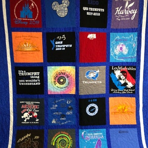 May include: A blue quilt with a white border, made from various t-shirts. The quilt is divided into 16 squares, each with a different design. Some of the designs include a Disney castle, a trumpet, a musical note, and a skull and crossbones. The text on the quilt includes "Disney 2019", "QHS Trumpets", "It's a Trumpet thing", "Arsenic and Old Lace", "Octoberfest Parade and Field", and "Cinderella".