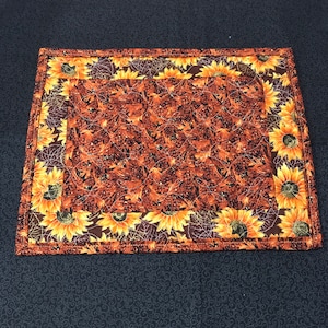 May include: A brown and yellow quilted placemat with a sunflower pattern. The placemat has a brown border with a white inner border.