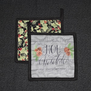 May include: Two quilted pot holders with a black and white border. The top pot holder has a green and red holly pattern. The bottom pot holder has a gray background with the text "Old Fashioned Hot Chocolate rich and creamy".