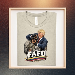 Puede incluir: Camiseta gris claro con un gráfico de dos hombres abrazándose. Uno lleva un traje, el otro una chaqueta y gafas de sol. El texto "FAFO" está debajo, con una cinta con los colores de la bandera colombiana.