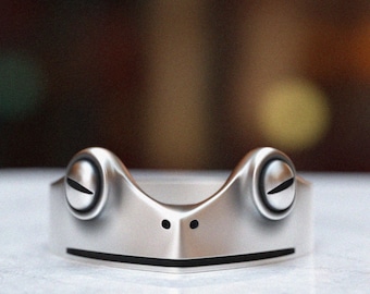 May include: A silver frog ring with a smooth, rounded surface and two large, round eyes. The ring is designed to look like a frog's head.
