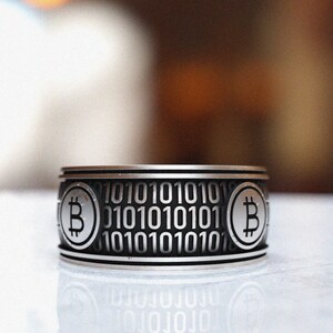 May include: A silver ring with a black band featuring a repeating pattern of the binary code "10101010" and a Bitcoin symbol on each side.