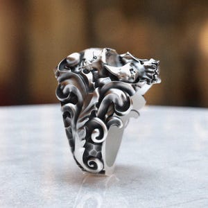 May include: A silver skull ring with intricate floral details. The skull is detailed with a textured surface and the ring band is smooth.