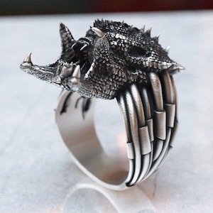 May include: A silver ring with a detailed, textured design of a dragon's head with an open mouth. The ring band is made of multiple bands of metal.