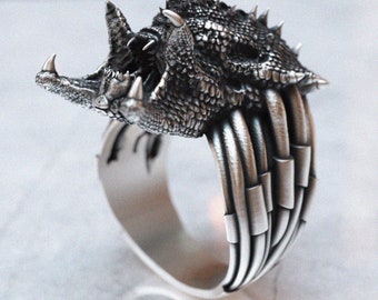 May include: A silver ring with a detailed, textured design of a dragon's head with an open mouth. The ring band is made of multiple bands of metal.