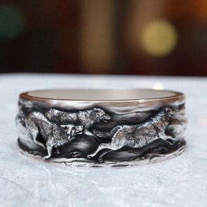 May include: A silver ring with a detailed design of three wolves running in a pack. The wolves are depicted in a realistic style with fur and muscle definition.