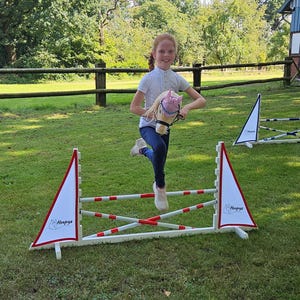 May include: A young person jumps over a white and red obstacle course with a white stuffed horse. The obstacle course has a white triangular sign with the word "Hoopy's" on it.