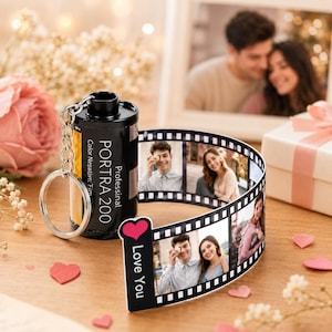Może przedstawiać: Czarny brelok w kształcie rolki filmu fotograficznego z wysuwanym paskiem zawierającym kilka wydrukowanych zdjęć pary. Pojemnik na film jest oznaczony jako Professional PORTRA 200. Na końcu znajduje się czerwone serce i napis Love You.