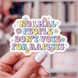 Puede incluir: Una pegatina colorida con el texto "NORMAL PEOPLE DON'T VOTE FOR RAPISTS" en una fuente retro. La pegatina tiene un borde blanco y está decorada con pequeños gráficos de flores y estrellas. El fondo es una imagen borrosa de un sofá rosa y una estantería.