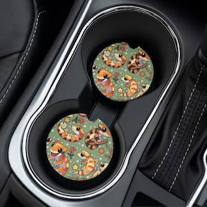 Puede incluir: Dos posavasos para coche con un patrón repetido de geckos de dibujos animados en naranja, marrón y amarillo sobre un fondo verde. Los posavasos están en los portavasos de un coche. Los geckos están rodeados de flores y estrellas.
