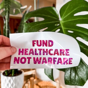 Pode incluir: Um autocolante branco com o texto "FUND HEALTHCARE NOT WARFARE" em letras cor-de-rosa brilhantes. O autocolante é segurado em frente de uma grande folha verde e outras plantas em vasos brancos.