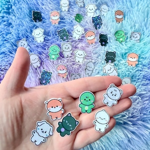 May include: A hand holding a collection of small, colorful stickers. The stickers feature various cartoon animals, including bears, cats, and dogs, in shades of orange, green, black, and white. The stickers are on a fluffy, blue and purple background.