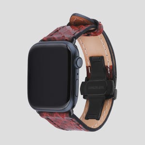 Könnte beinhalten: Eine dunkelblaue Smartwatch mit einem roten Lederarmband mit Schlangenmuster. Das Armband hat einen schwarzen Edelstahlverschluss. Das Zifferblatt der Uhr ist schwarz.