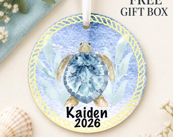 May include: A round ceramic ornament featuring a watercolor sea turtle illustration on a blue textured background, framed by a yellow rope border. The text Kaiden 2026 is printed in black below the turtle. A white ribbon is attached at the top.