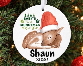 May include: A round white ceramic Christmas ornament featuring a watercolor illustration of a sleeping fawn wearing a red Santa hat. The text reads Baby's First Christmas, the name Shaun, and the year 2026. It hangs from a white ribbon.