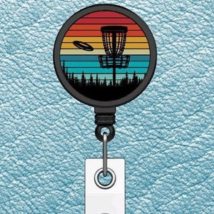 May include: A black retractable badge holder with a round, colorful disc featuring a silhouette of a disc golf basket and a flying disc against a sunset background.