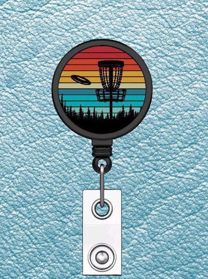 May include: A black retractable badge holder with a round, colorful disc featuring a silhouette of a disc golf basket and a flying disc against a sunset background.