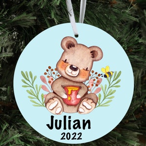 May include: A blue and white Christmas ornament featuring a cartoon bear holding a pot of honey. The ornament has the name "Julian" and the year "2022" printed on it.