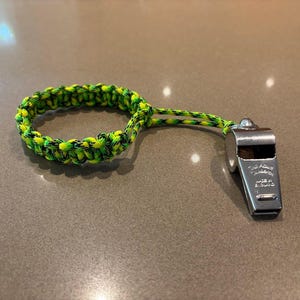 May include: A silver whistle with a green and yellow braided lanyard. The whistle is made of metal and has text on the side. The lanyard is a loop of braided cord.