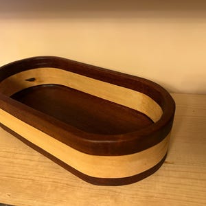 Puede incluir: Cuenco ovalado de madera con borde y base marrón oscuro, y una banda marrón claro en el medio. El interior es marrón oscuro. El cuenco está sobre una superficie de madera marrón claro.