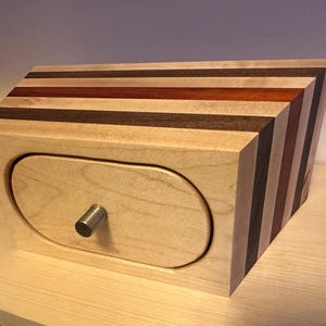 May include: A wooden box with a light-colored front and a drawer with a metal knob. The top and side feature a striped design with various wood tones, including light, dark, and reddish-brown.