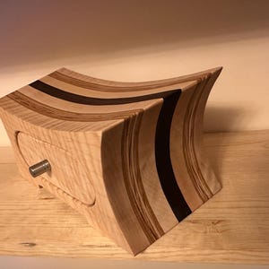 May include: A wooden box with a curved design and a drawer. The box is made of light wood with dark stripes and a black accent. The drawer has a metal knob.