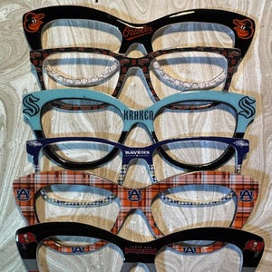 Puede incluir: Cinco pares de gafas de ojo de gato de colores con diferentes logotipos de equipo. Los logotipos incluyen los Baltimore Orioles, los Seattle Kraken, los Baltimore Ravens, los Auburn Tigers y los Tampa Bay Buccaneers.