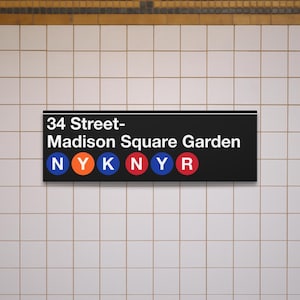 May include: A black rectangular sign with white text reading "34 Street-Madison Square Garden." Below the text are colored circles with the letters NYKNYR. The sign is mounted on a tiled wall.