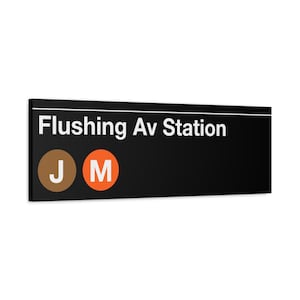 May include: Black and white subway station sign with the text "Flushing Av Station" in white. The sign also has two circles with the letters "J" and "M" in white, the "J" circle is brown and the "M" circle is orange.