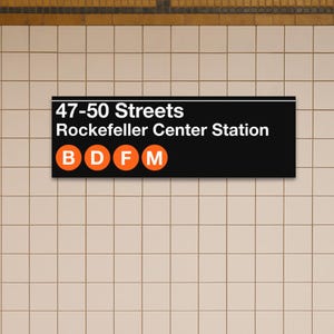 May include: A rectangular black subway station sign with white text reading 47-50 Streets Rockefeller Center Station, featuring four orange circular icons containing the white letters B, D, F, and M, mounted on a wall of white square tiles.