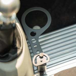 May include: A black metal car accessory with a circular top and a series of holes along its arm. The accessory is mounted on a silver, ridged surface, near a gear shift and a black interior.