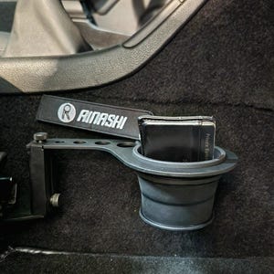 May include: Black car cup holder with a black wallet inside. The cup holder is attached to the side of the car with a metal bracket. The cup holder has a black strap with the text "Rimashi" on it.