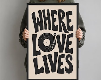 May include: A rectangular framed wall art print featuring the black text WHERE LOVE LIVES on a light beige background. The letter O in the word LOVE is replaced by a black vinyl record graphic with a tone arm, all enclosed in a thin black frame.