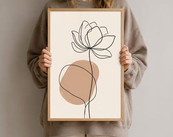 Könnte beinhalten: Ein gerahmter vertikaler Wandkunstdruck mit einer durchgehenden schwarzen Linienzeichnung einer Lotusblume, die auf einem cremefarbenen Hintergrund zentriert ist. Hinter dem Blumenstiel bildet eine feste, abgerundete, hellbraune Form einen Kontrast.