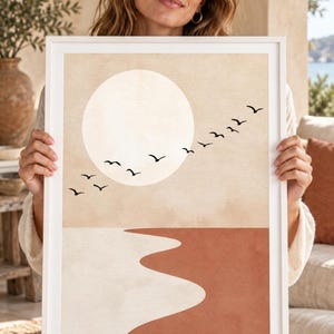 Könnte beinhalten: Ein gerahmter Kunstdruck mit einer großen, blassbeigen Sonne vor einem hellbraunen Himmel. Ein Schwarm schwarzer Vögel fliegt über die Sonne. Der untere Teil des Bildes zeigt eine wellenförmige, abstrakte Landschaft mit einem cremefarbenen und einem terrakottafarbenen Bereich.