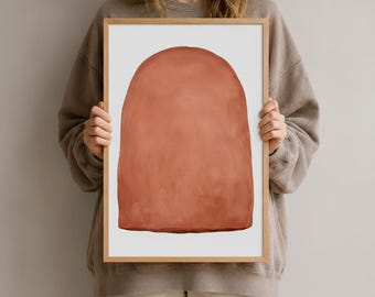 Könnte beinhalten: Ein gerahmter Kunstdruck mit einer einfarbigen, terrakottafarbenen Bogenform, die mittig auf einem schlichten weißen Hintergrund platziert ist. Das Kunstwerk befindet sich in einem hellen Holzrahmen und zeigt eine klare, einfache Komposition.
