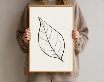 Könnte beinhalten: Ein gerahmter vertikaler Wandkunstdruck mit einer schwarzen Tuschezeichnung eines einzelnen Blattes, das auf einem cremefarbenen Hintergrund zentriert ist. Das Kunstwerk befindet sich in einem hellen Holzrahmen mit schlichtem Rand.