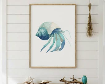 May include: A framed watercolor print of a hermit crab. The artwork features a blue and teal crab with a spiraled shell. The crab is set against a white background, and the frame is a light brown color. The artwork is displayed on a white wall.