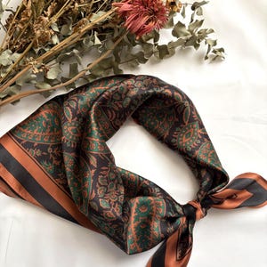 Op de afbeelding: Een vierkante zijden sjaal met een donkerbruine basis en een ingewikkeld paisley-patroon in tinten groen en koper. De sjaal heeft een brede koperkleurige rand langs de zijkanten en is in een losse lus geknoopt, met een gladde, satijnachtige afwerking.